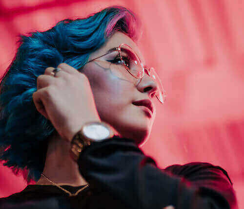 Person with blue hair and glasses, wearing a black top, against a pink background.
