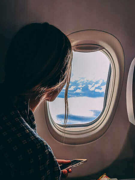 A woman with long hair gazes out of an airplane window at blue skies and clouds.