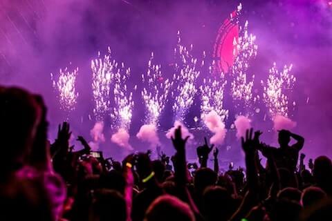 Crowd with raised hands, vibrant purple lights, and white fireworks in the background.