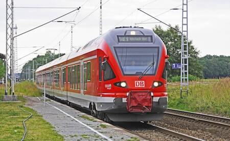Red and silver train on tracks, surrounded by green fields and cloudy sky.