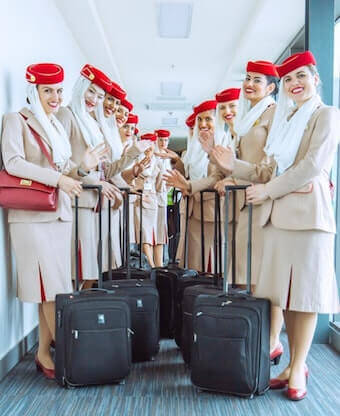 A group of flight attendants in beige uniforms and red hats, with black suitcases.