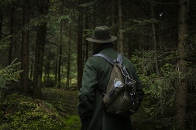 A person in a green coat and hat walks through a dark forest.