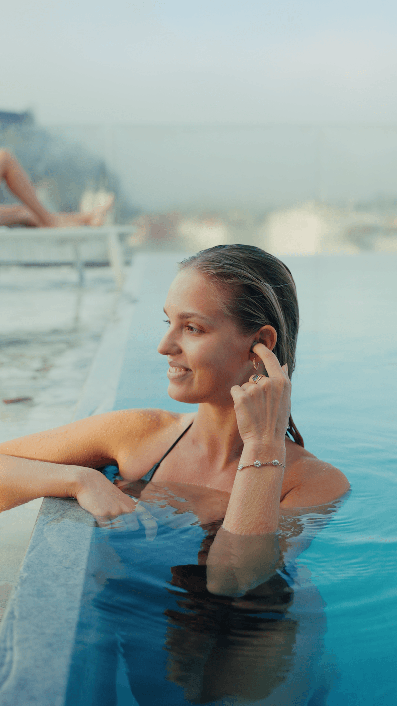 Alt text: Woman relaxing in an outdoor pool touches the earplug in her ear while smiling, with calm blue water and a softly blurred spa or rooftop setting in the background.