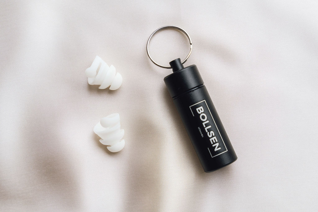Black container with "BOLL-SEN" label, two white earplugs on light fabric background.