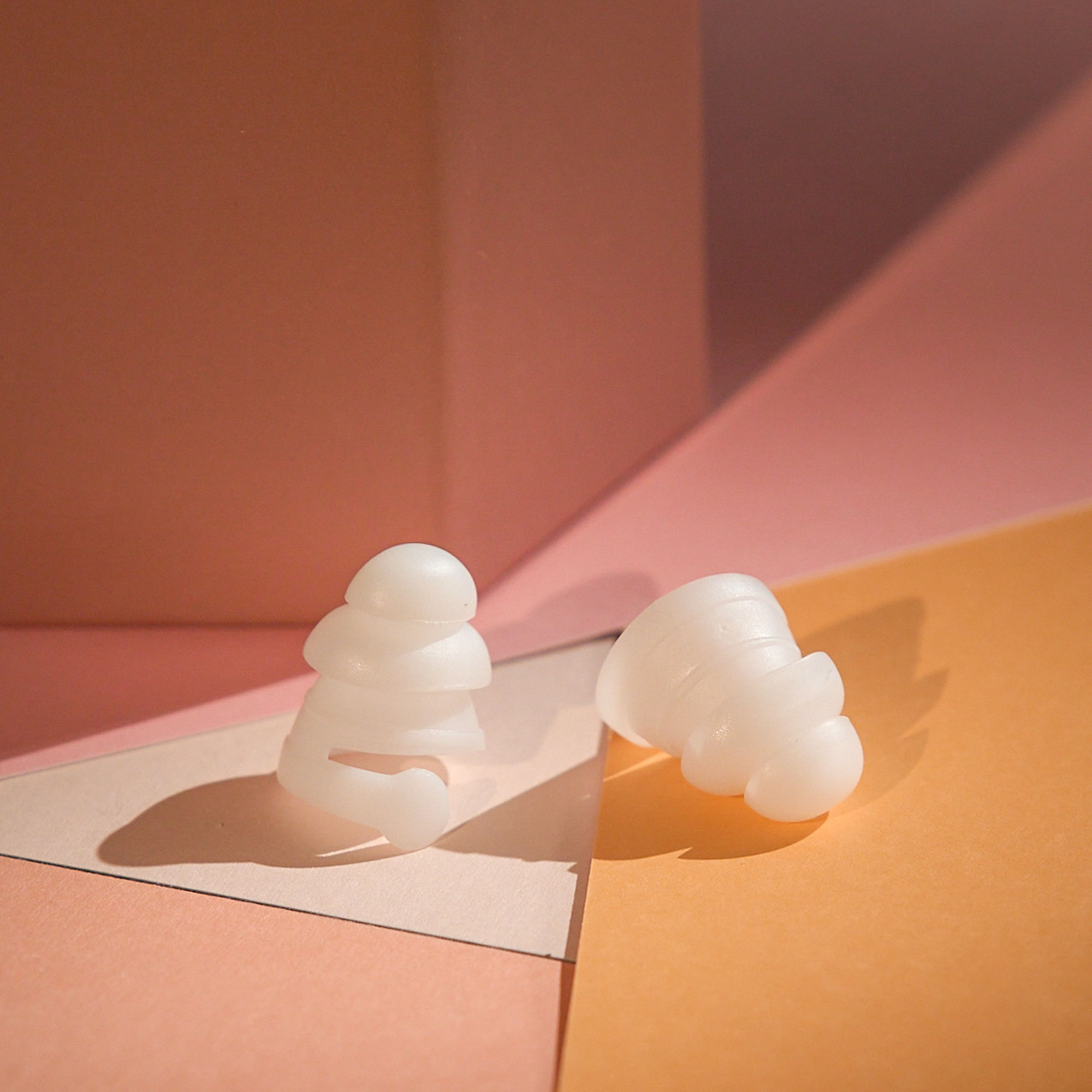 Two white earplugs on pastel geometric backgrounds of pink and orange.