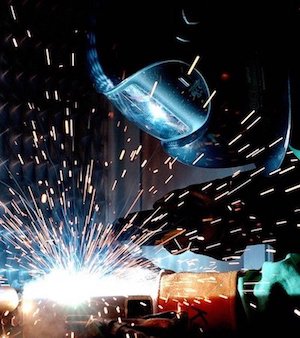 A welder in a blue helmet creates bright sparks against a dark background.