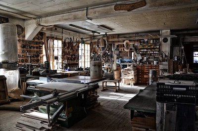 A cluttered workshop with wooden tables, tools, and large windows letting in natural light.