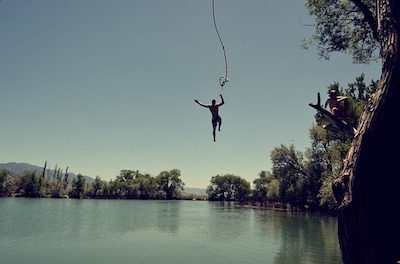 A person swings from a rope over a calm, green lake under a clear sky.