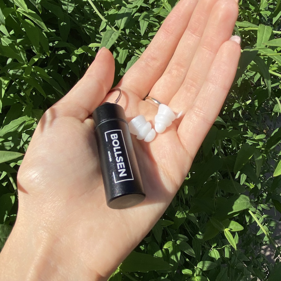 A hand holding a black container and white BOLLSEN Earplugs Hearing Protection against green foliage.