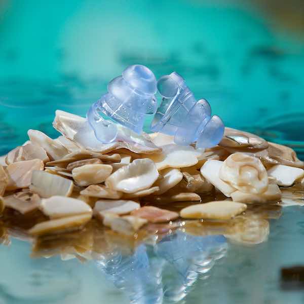 Two clear blue earplugs rest on a bed of seashells. Soft turquoise background.