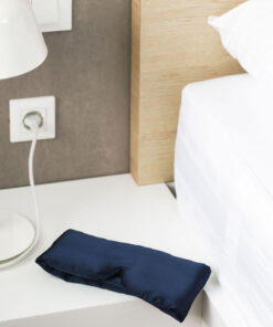 Navy blue sleep mask on a white bedside table next to a lamp and bed.
