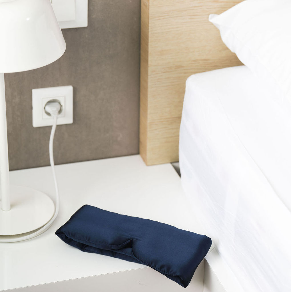 Navy blue sleep mask on a white bedside table next to a lamp and bed.
