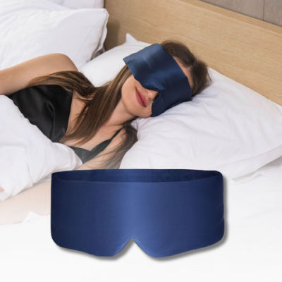 Woman resting on a bed, wearing a blue satin eye mask. Soft white pillows nearby.