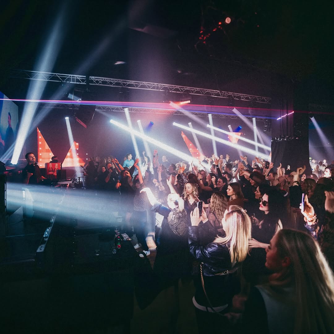 Crowd dancing under bright lights, with a DJ and vibrant laser beams.