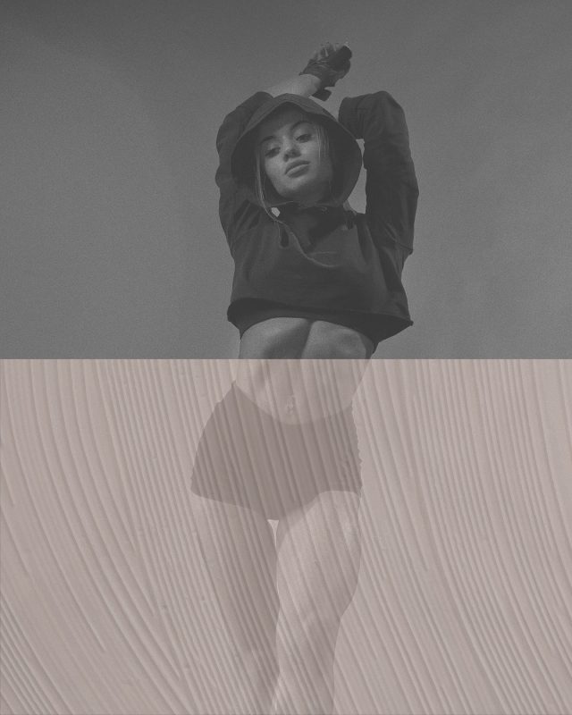 A woman in a black hoodie and shorts poses against a textured, soft pink background.