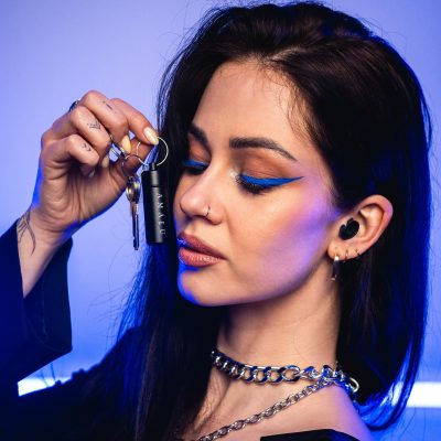 A woman with blue eyeliner holds black keys near her face, under soft blue lighting.