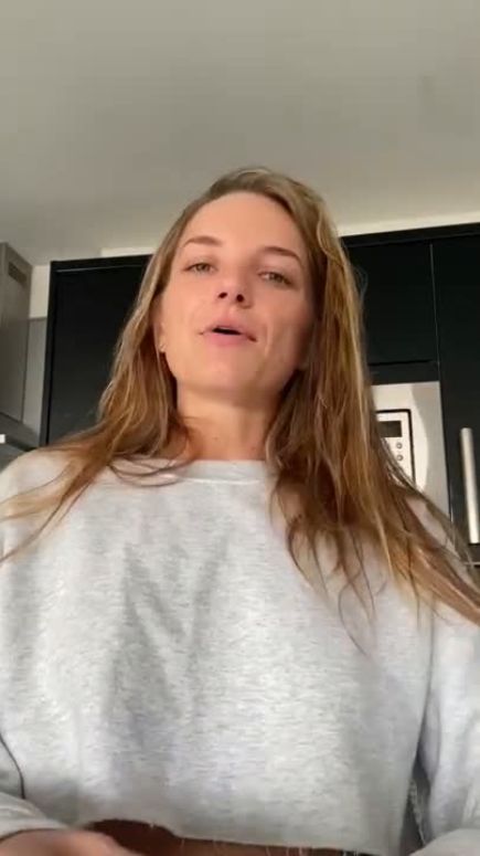 A woman with long hair wearing a grey sweater stands in a modern kitchen.