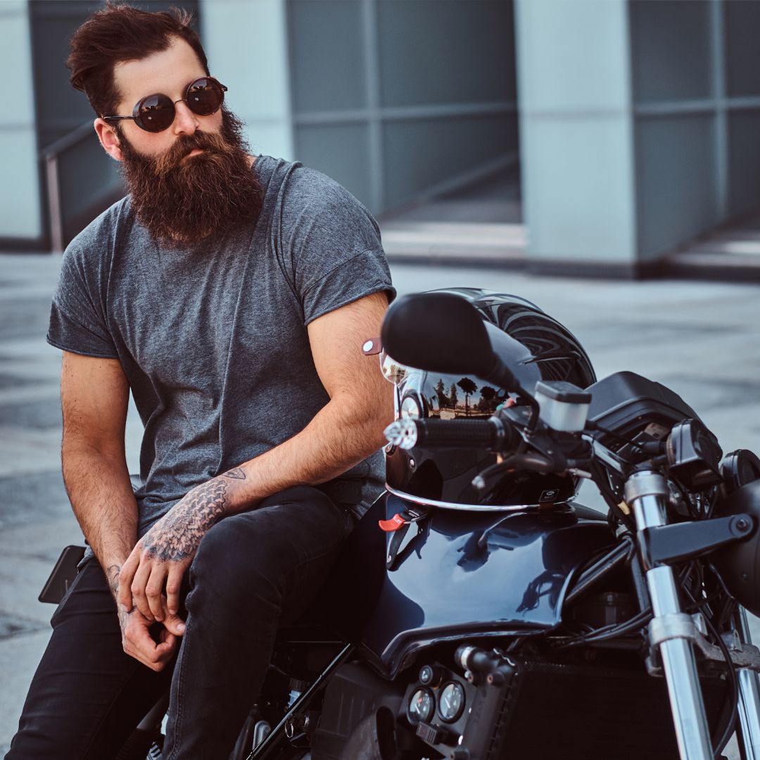 Man with a beard and sunglasses sitting on a black motorcycle, wearing a grey t-shirt.