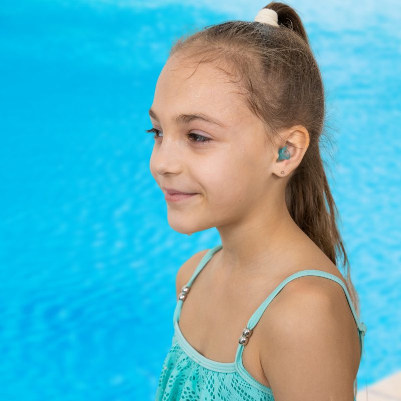 Young girl with brown hair, wearing a turquoise swimsuit, smiles by a blue pool.