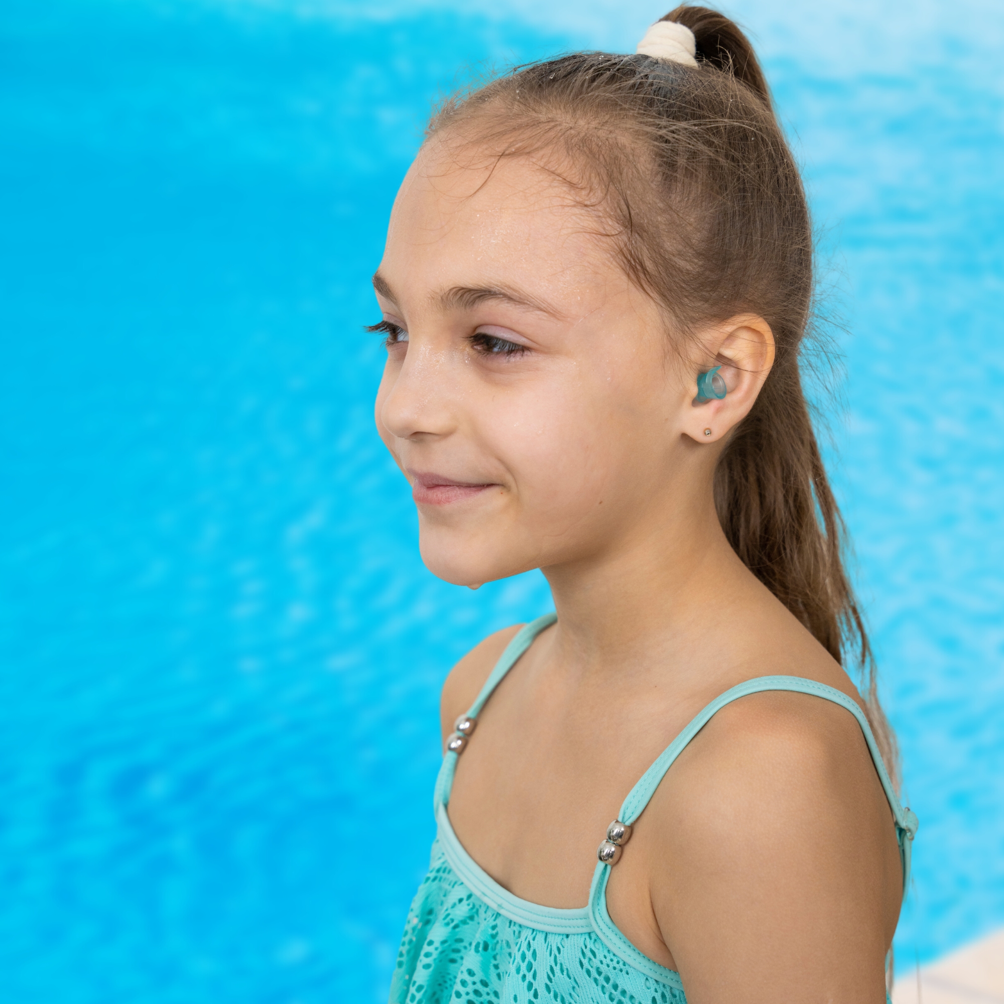 Young girl with brown hair, wearing a turquoise swimsuit, smiles by a blue pool.