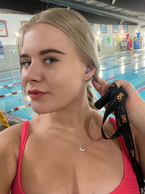 Young woman with blonde hair, wearing a pink swimsuit, near a swimming pool.