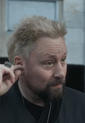Man with short, light blonde hair, wearing a black outfit, touching his ear.