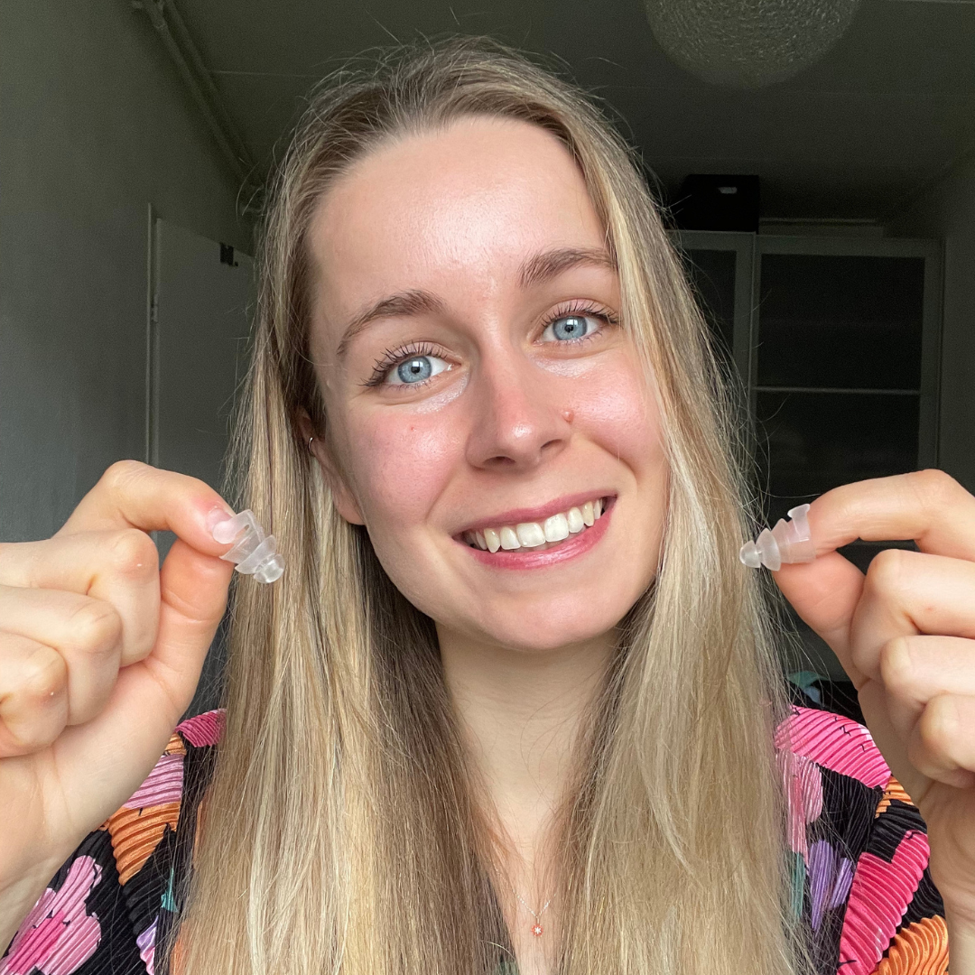 Smiling woman with long blonde hair holding clear earplugs, wearing a colourful patterned top.