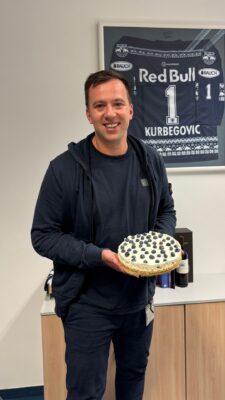 Man in navy hoodie holding a cheesecake topped with blueberries, office background. Founder of BOLLSEN Hearing Protecion