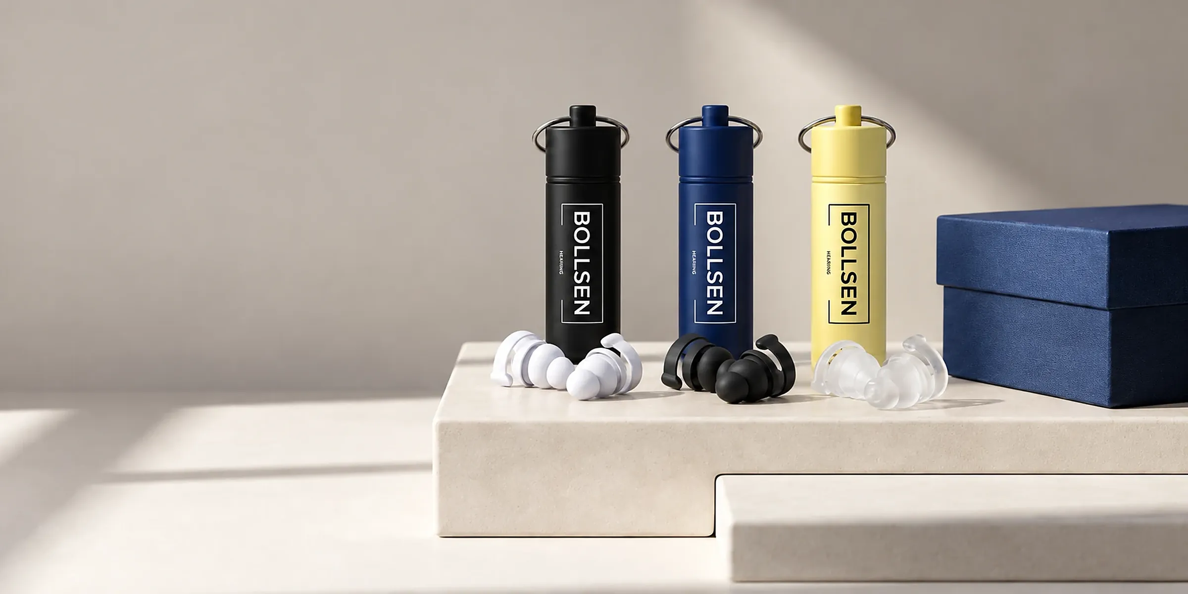 BOLLSEN earplug cases in black, navy and yellow with white, black and clear earplugs and a navy gift box on a stone plinth